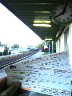 Train tickets saying departure is 6:10am held in front of a clock that reads 8:05 while an oncoming train arives in the distance.  Greve.