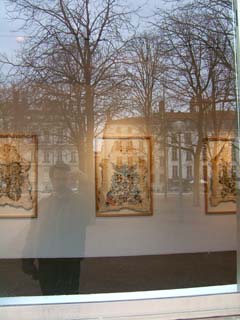 A view through the window into Le Rectangle at three picture frames holding decorated animal skins.  Reflectgted in the glass is my image holding a small camera and the winter scene of Lyon in the background.